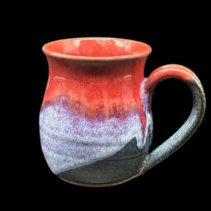 May include: Handmade ceramic mug with a vibrant color gradient. The top is a rich red, transitioning through purple and blue to a dark base. The handle is blue with dark accents. A unique piece for coffee or tea.