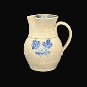 Puede incluir: Una jarra de gres con cuerpo color crema y diseño floral azul. La jarra tiene asa y pico vertedor. La altura es de aproximadamente 23 cm.