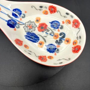 May include: White ceramic spoon rest with a red rim and floral design. The spoon rest features red, blue, and orange flowers with green stems. The spoon rest is designed to hold a spoon while cooking.