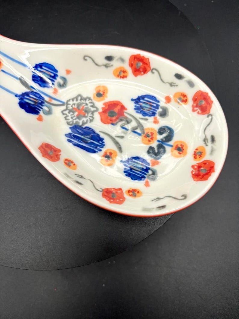 May include: White ceramic spoon rest with a red rim and floral design. The spoon rest features red, blue, and orange flowers with green stems. The spoon rest is designed to hold a spoon while cooking.