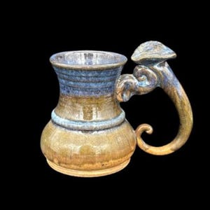 May include: A handcrafted ceramic mug with a distinctive mushroom-shaped handle. The mug has a rounded base and a flared rim, with a colour scheme of brown, blue, and gold. The handle is intricately designed, enhancing the mug's artistic appeal.
