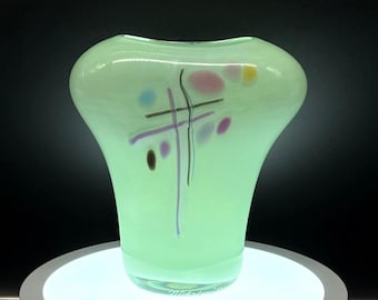 May include: A pale green glass vase with a bulbous body and a flared top. The vase features abstract markings in purple, pink, and yellow. The vase is displayed on a white, circular platform. The vase's height is approximately 20 cm.