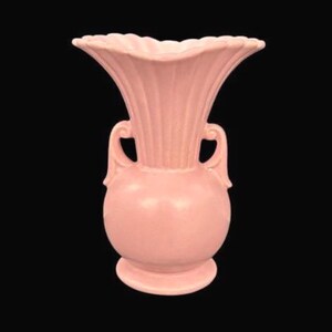 May include: A vintage pink ceramic vase with a flared, ruffled rim and two curved handles. The vase has a bulbous body and a pedestal base. The exterior features vertical ribbed detailing. The background is black.