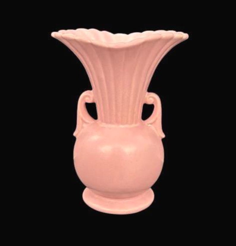 May include: A vintage pink ceramic vase with a flared, ruffled rim and two curved handles. The vase has a bulbous body and a pedestal base. The exterior features vertical ribbed detailing. The background is black.