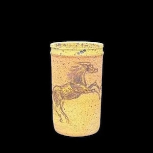 Op de afbeelding: Een cilindrische, handgemaakte keramische beker met een getextureerde, gespikkelde geel-bruine afwerking. Een bruin paard in een steigerende pose is afgebeeld op de zijkant. De rand is donkerder geel.