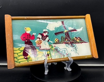 May include: A vintage framed print featuring a Dutch scene with a windmill, figures in traditional attire, and a basket. The artwork has a light blue background and a wooden frame. The print is displayed on a clear stand.