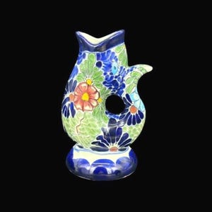 May include: A hand-painted ceramic pitcher with a unique design. The pitcher features a vibrant floral pattern in shades of blue, green, red, and yellow. It has a distinctive handle and a circular opening in the body. The base is a rounded shape.