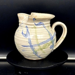 May include: A handmade ceramic pitcher with a cream-colored base and a curved handle. The pitcher is decorated with blue and green abstract brushstrokes. The spout is slightly angled for easy pouring. The pitcher is a unique piece of pottery.