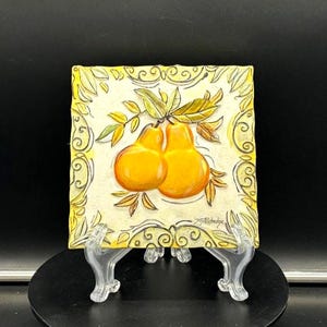 May include: A square, hand-painted ceramic tile featuring two ripe, orange pears with green and brown leaves. The tile has a yellow border with decorative swirls and the artist's signature.
