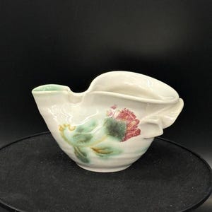 Op de afbeelding: Een witte keramische creamer met een bloemenontwerp in rood, groen en geel. De creamer heeft een unieke, asymmetrische vorm met een tuit en een handvatachtige verlenging. Het bloemmotief is handgeschilderd.