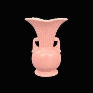 May include: A light pink ceramic vase with a fluted, ruffled top and two curved handles. The vase has a bulbous body and a circular base, standing against a black background. This decorative vase is suitable for floral arrangements.