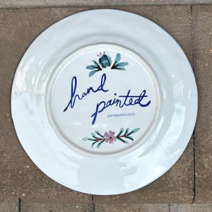 以下が含まれることがあります： 手描きのデザインが施された白いセラミックプレート。中央には、青い筆記体で「hand painted」の文字があり、ティールとピンクの花柄のアクセントが付いています。「ANTHROPOLOGIE」の文字も表示されています。