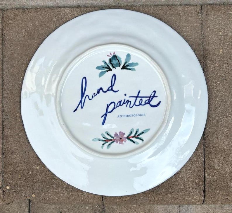 以下が含まれることがあります： 手描きのデザインが施された白いセラミックプレート。中央には、青い筆記体で「hand painted」の文字があり、ティールとピンクの花柄のアクセントが付いています。「ANTHROPOLOGIE」の文字も表示されています。