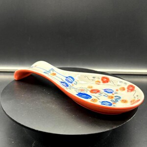 May include: A ceramic spoon rest with a curved handle and a wide, oval bowl. The spoon rest is decorated with blue and red floral patterns on a cream background, with an orange rim.