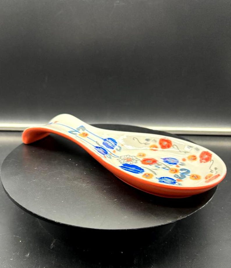 May include: A ceramic spoon rest with a curved handle and a wide, oval bowl. The spoon rest is decorated with blue and red floral patterns on a cream background, with an orange rim.