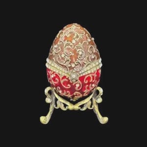 May include: An ornate, jeweled egg on a decorative stand. The egg features a red base with a swirling pattern and a brown top with intricate filigree and sparkling accents. The stand is gold-toned with curved legs.