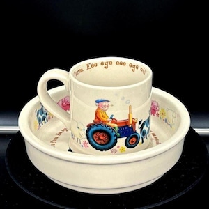 May include: Cream-colored ceramic mug and bowl set with a farm theme. The mug features a farmer driving a red and blue tractor. The bowl and mug are decorated with farm animals and the phrase "Eee aye eee aye oh!".