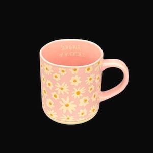 May include: A light pink ceramic mug featuring a repeating pattern of white daisies with yellow centers. The interior rim of the mug displays the text "bonjour, mon amour" in a simple, light-colored font. The mug has a curved handle on the right side.