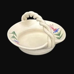 Pode incluir: Cesto de cerâmica creme com uma alça torcida. O cesto apresenta um design floral com detalhes em azul, rosa e verde. A alça tem um design espesso em forma de corda. O cesto está sobre um fundo preto.
