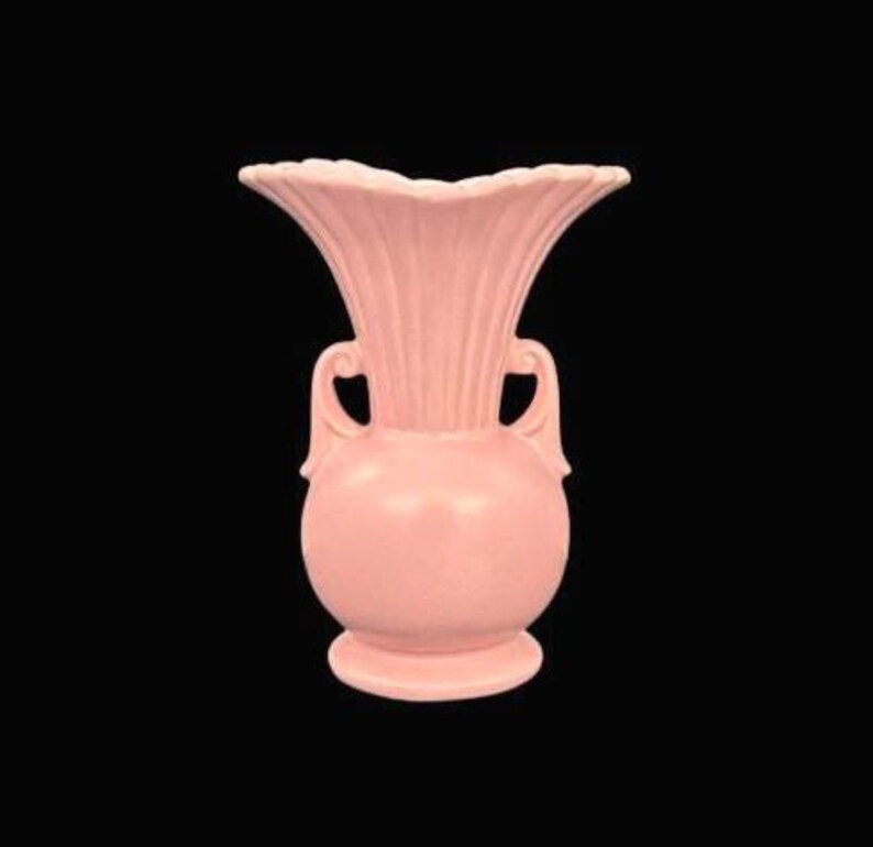 May include: A pink ceramic vase with a flared, ruffled top and two curved handles. The vase has a bulbous body and a round base. The fluted design on the top adds a decorative touch. The vase is a soft, muted pink color.
