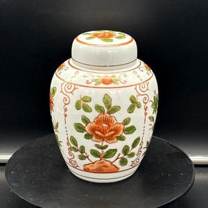Puede incluir: Un tarro de jengibre de cerámica blanca con tapa, adornado con motivos florales naranjas y verdes. El tarro tiene forma redondeada y está decorado con flores y follaje estilizados. La tapa refleja el diseño floral.