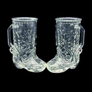 May include: Two clear glass boot-shaped mugs with handles. The mugs have a detailed floral and vine pattern. The boots are standing upright and are side by side. The background is black.