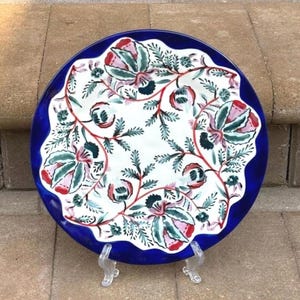 以下が含まれることがあります： 赤、ピンク、緑、青の花柄が施された白い中心部を持つ丸いセラミックプレート。プレートは波状の濃い青色の縁があり、透明なプラスチック製のスタンドに展示されています。花柄には、様式化された花と葉が含まれています。