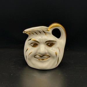May include: A small white ceramic pitcher with a gold-painted face and handle. The face has a whimsical expression with a wide smile and gold-painted details.