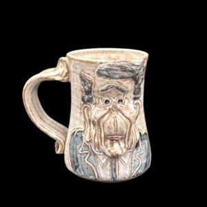 May include: A beige ceramic mug with a sculpted face design. The face has glasses, a mustache, and a suit. The handle is curved and the mug has a textured surface. The mug is a unique piece of art.