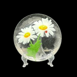 Puede incluir: Un plato de vidrio transparente con un diseño de margaritas pintadas. Las margaritas tienen pétalos blancos, centros amarillos y una pequeña mariquita roja. El plato se exhibe sobre un soporte decorativo transparente.