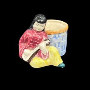 Könnte beinhalten: Keramikfigur einer Frau, die ein Saiteninstrument spielt, mit rotem Oberteil und gelber Hose. Daneben steht ein blau-weißer Korb. Die Figur hat schwarzes Haar und ist vor schwarzem Hintergrund.