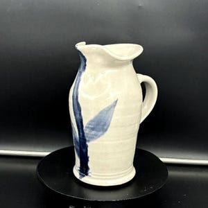 Puede incluir: Jarra de cerámica hecha a mano con base color crema y diseño floral azul. La jarra tiene un asa curva y un pico. El diseño azul es una hoja y un tallo estilizados. La jarra mide aproximadamente 20 cm de altura.
