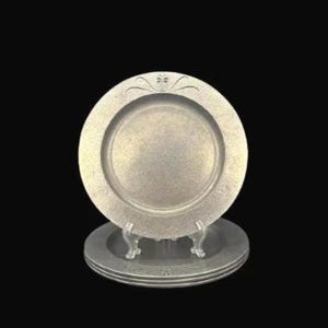 Puede incluir: Platos de peltre apilados con un diseño floral decorativo. Los platos se exhiben sobre un soporte transparente sobre un fondo negro. Los platos tienen un borde ligeramente elevado y un área central empotrada.