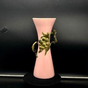 May include: A pink vase with a flared top and a narrow base. The vase features a gold-coloured grape cluster and vine design on the front and handles. The vase is approximately 20 cm tall.