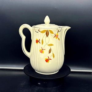 May include: Cream-colored vintage pitcher with a lid, handle, and spout. The pitcher is decorated with a floral design in shades of yellow, orange, and brown. The rim and handle are trimmed with gold. The pitcher is sitting on a black surface.
