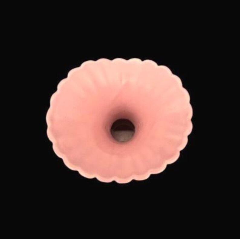 May include: A light pink, donut-shaped vase with a scalloped edge. The vase has a central opening and a smooth, matte finish. The background is solid black.