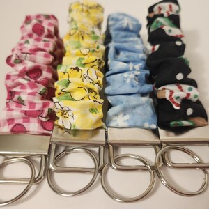 May include: Four fabric keychains with metal hardware. The fabric patterns include pink gingham with strawberries, yellow floral, blue snowflakes, and black with Christmas designs. Each keychain has a silver metal rectangular plate and a circular metal loop.