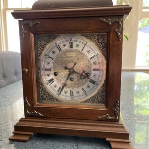 May include: A wooden mantel clock with a brass handle on top and a silver face with Roman numerals. The clock has a decorative design around the face and a dark wood finish.