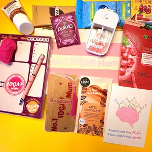 May include: A selection of self-care products on a yellow surface. Items include a pink planner, pen, bookmark, face mask, lip balm, hand cream, tea, wipes, and a spray bottle with the text "No.1 IDGAF Mum".