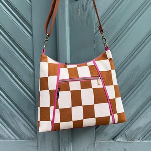 Könnte beinhalten: Eine braun-weiß karierte Umhängetasche mit pinkfarbenem Reißverschluss und Besatz. Die Tasche hat einen braunen Riemen und hängt vor einer türkisfarbenen Tür.