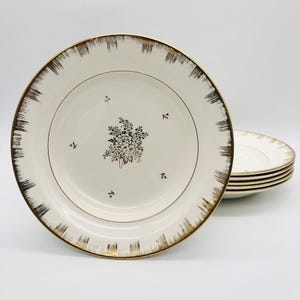 May include: A set of vintage cream-coloured plates with a gold rim and a floral centre design. The plates have a gold trim with a brushstroke effect. A stack of plates is visible in the background.