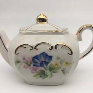 Può includere: Una teiera bianca con un bordo dorato e un motivo floreale. La teiera ha un coperchio e un manico. I fiori sono blu, rosa e gialli.