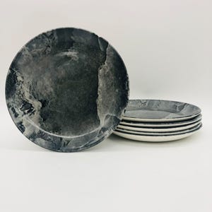 Puede incluir: Un juego de seis platos de cerámica con un diseño veteado gris y blanco. Los platos tienen un borde gris oscuro y una parte inferior blanca. Los platos son redondos y tienen una estética moderna.