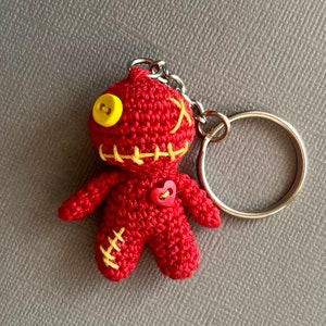 May include: Red crocheted doll keychain with yellow button eyes, yellow stitching, and a red heart button. The doll has a stitched-on smile and is attached to a silver key ring.
