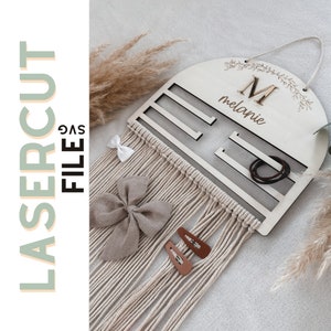 Op de afbeelding: Een houten haaraccessoire-organizer met de naam "melanie" en de letter "M" gegraveerd. De organizer heeft sleuven voor haarbanden en clips, en een macrame franje. De tekst "LASERCUT FILE SVG" staat aan de linkerkant.