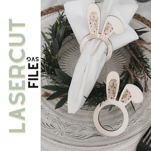 Op de afbeelding: Houten paashaas servetringen met bloemendetails op de oren en de tekst "Frohe Ostern". De ringen liggen op een heldere glazen plaat met een witte servet. De tekst "Laser Cut File SVG" is ook aanwezig.