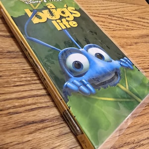May include: A Bugs Life VHS tape. The cover features a blue cartoon insect with large eyes and antennae. The title "a bug's life" is in yellow letters. The background is green with a leaf design. The tape is in a plastic sleeve.