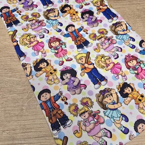Op de afbeelding: Witte stof bedekt met kleurrijke cartoonpoppen, elk met unieke outfits en kapsels. De poppen zijn verspreid over de stof, die ook is versierd met pastelkleurige stippen. De poppen bevinden zich in verschillende poses.