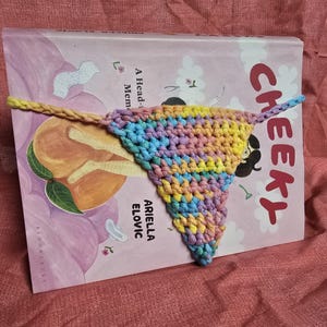 May include: A multi-colored crochet bookmark in the shape of a triangle is placed on a book titled "Cheeky" by Ariella Elovic. The book cover features a peach and a floral design.