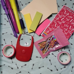 May include: A flat lay of various stationery supplies on a blue background with a white arrow pattern. The items include sticky notes, pens, a magnifying glass, a roll of tape, a small notebook, and a bag of paper clips.